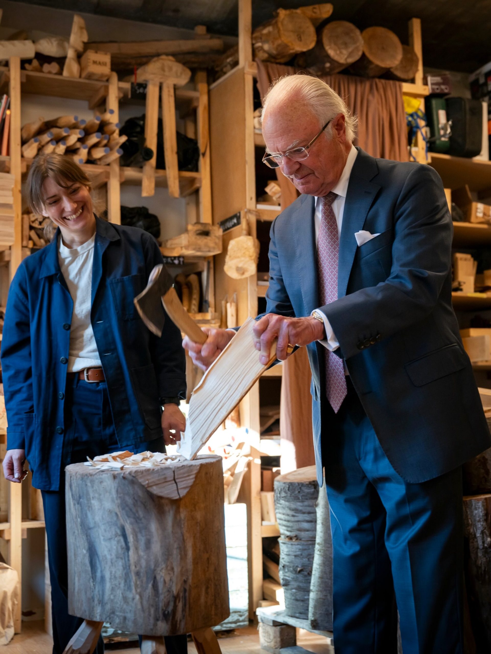 King Carl XVI Gustaf of Sweden, in a dark suit, splitting wood with an axe over a chopping block while axe maker Julia Kalthoff in a blue work jacket laughs beside him, with shelves of axe handles and timber behind them