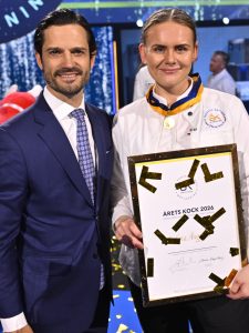 Evelina Anggren in a white chef jacket holding the framed Chef of the Year 2026 diploma beside Prince Carl Philip, gold confetti falling at Annexet in Stockholm