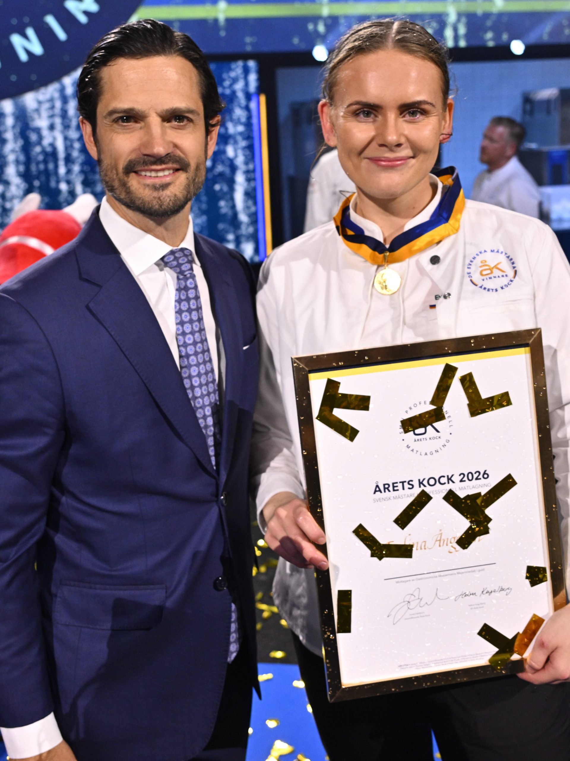 Evelina Anggren in a white chef jacket holding the framed Chef of the Year 2026 diploma beside Prince Carl Philip, gold confetti falling at Annexet in Stockholm