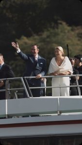 Grand Duke Guillaume of Luxembourg waving in a navy suit with a pink tie, Grand Duchess Stephanie smiling beside him in white, on the deck of the M.S. Princesse Marie-Astrid with green Moselle hills behind them