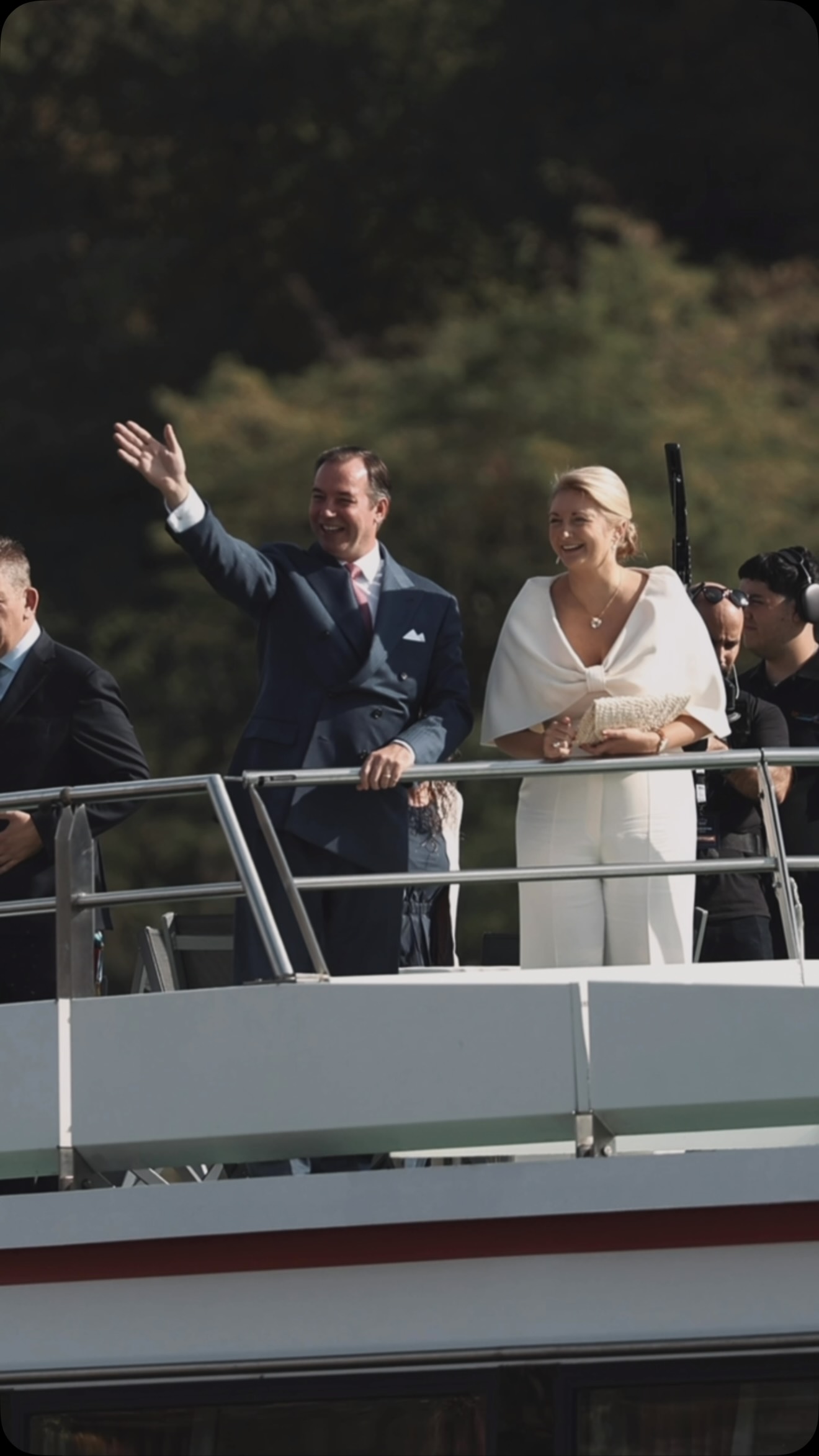 Grand Duke Guillaume of Luxembourg waving in a navy suit with a pink tie, Grand Duchess Stephanie smiling beside him in white, on the deck of the M.S. Princesse Marie-Astrid with green Moselle hills behind them