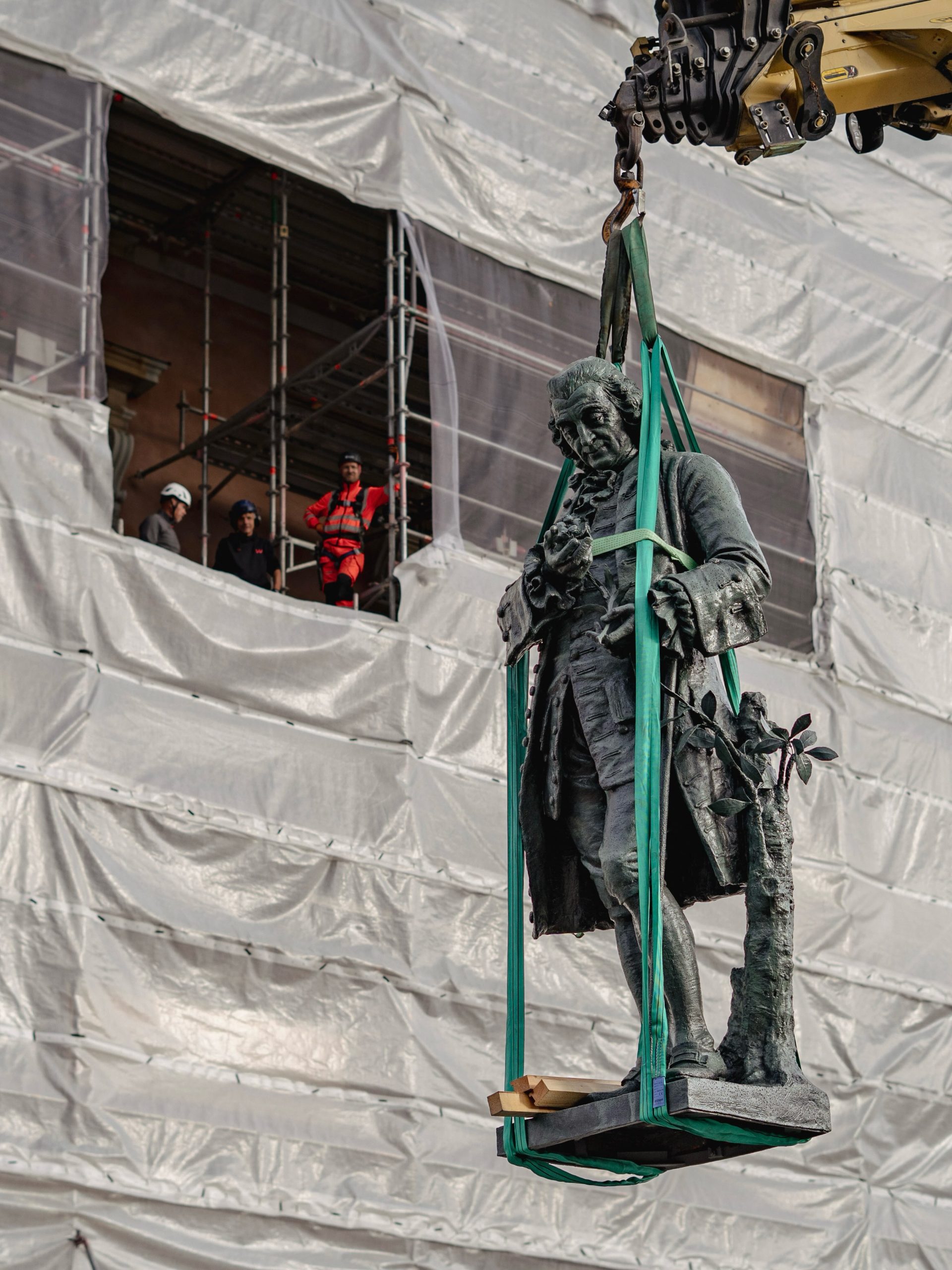 A restored dark bronze statue of Carl von Linne hangs in green crane straps before Stockholm's Royal Palace facade, wrapped in white sheeting with scaffolding and helmeted workers.